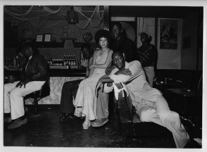 Black-and-white 1970s bar scene; friends lounging by the counter in a dim, stylish nightclub.