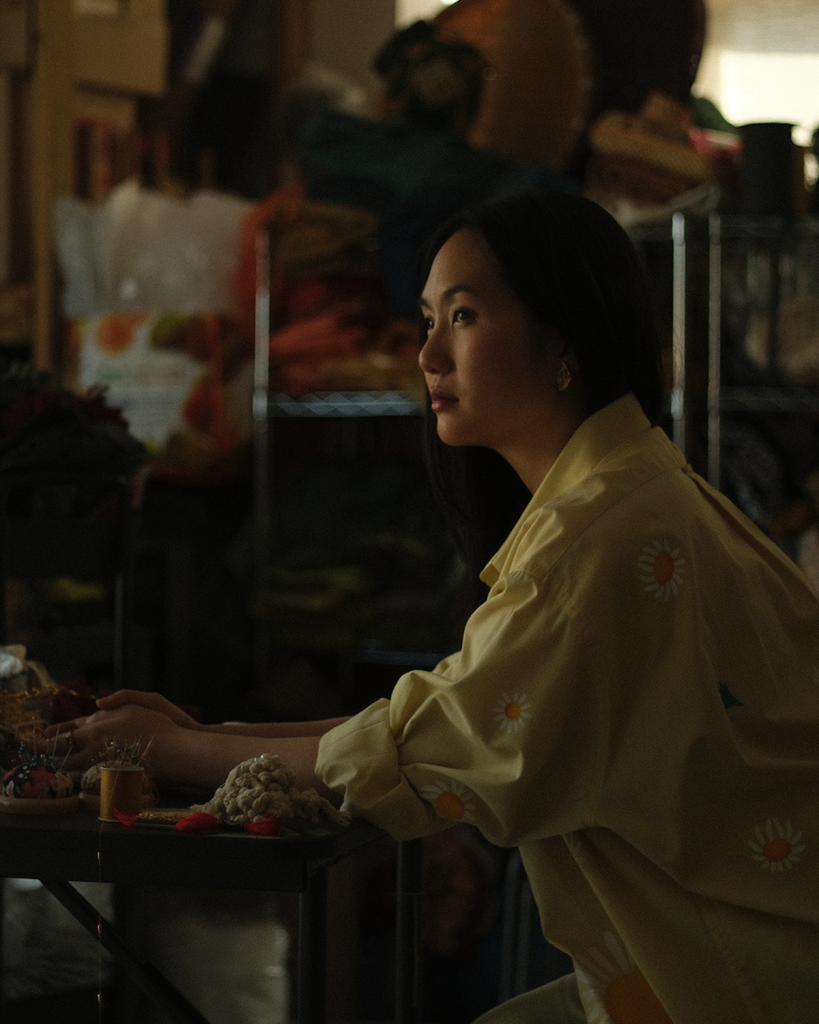 Woman in yellow daisy shirt working at craft table in dim studio, surrounded by fabrics and sewing supplies