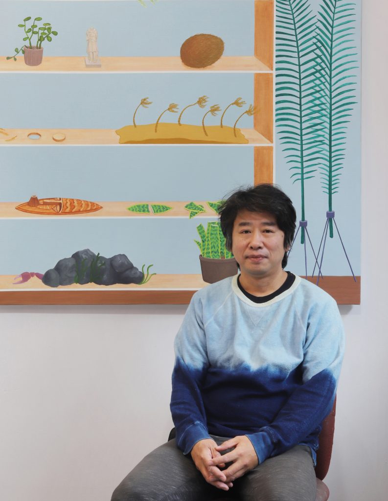 Artist seated in studio before colorful painting of plants, coconut, boat and rocks on blue shelves.