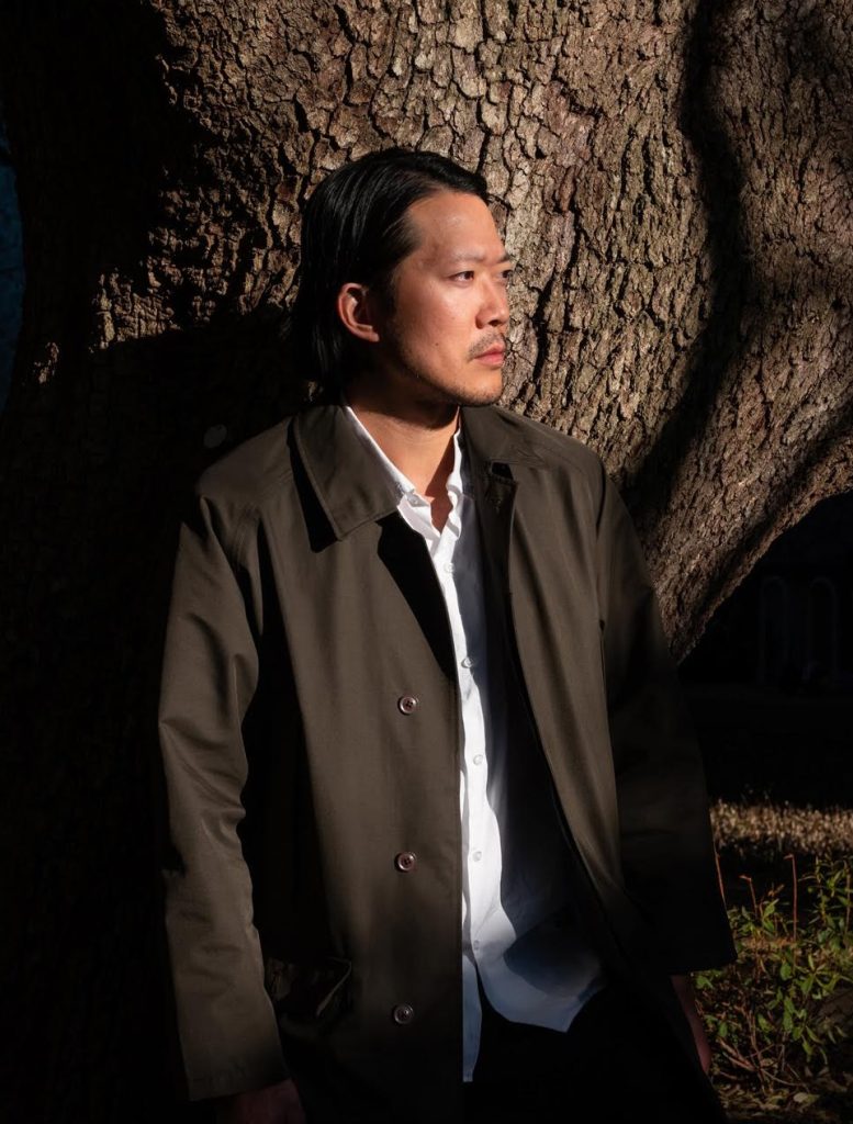 Thoughtful man in dark coat by a tree, side-lit outdoor portrait with textured bark and shadows in golden hour.