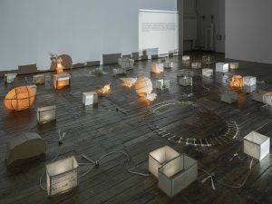 Contemporary mixed-media installation with glowing paper lanterns, cube lights, and projected text on a wooden gallery floor