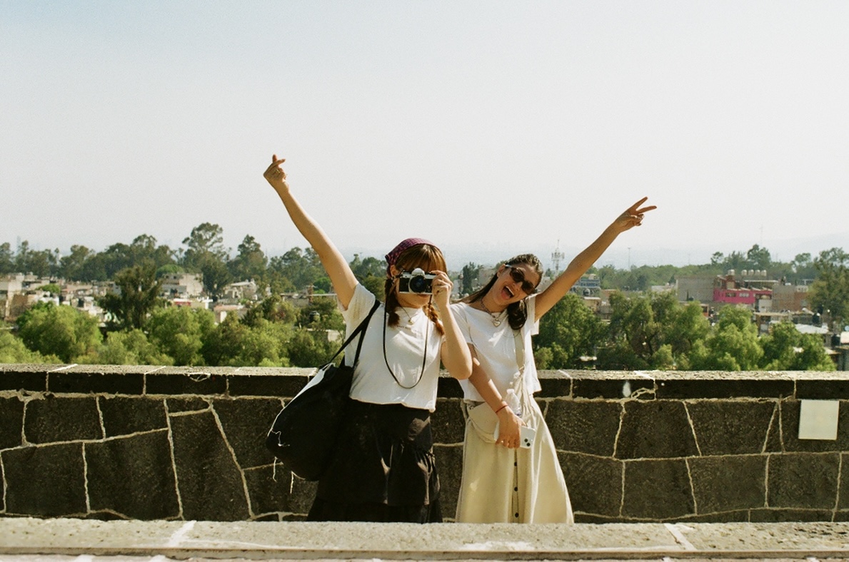 Two travelers posing with camera and peace signs on a stone terrace, overlooking a green cityscape on a sunny day.