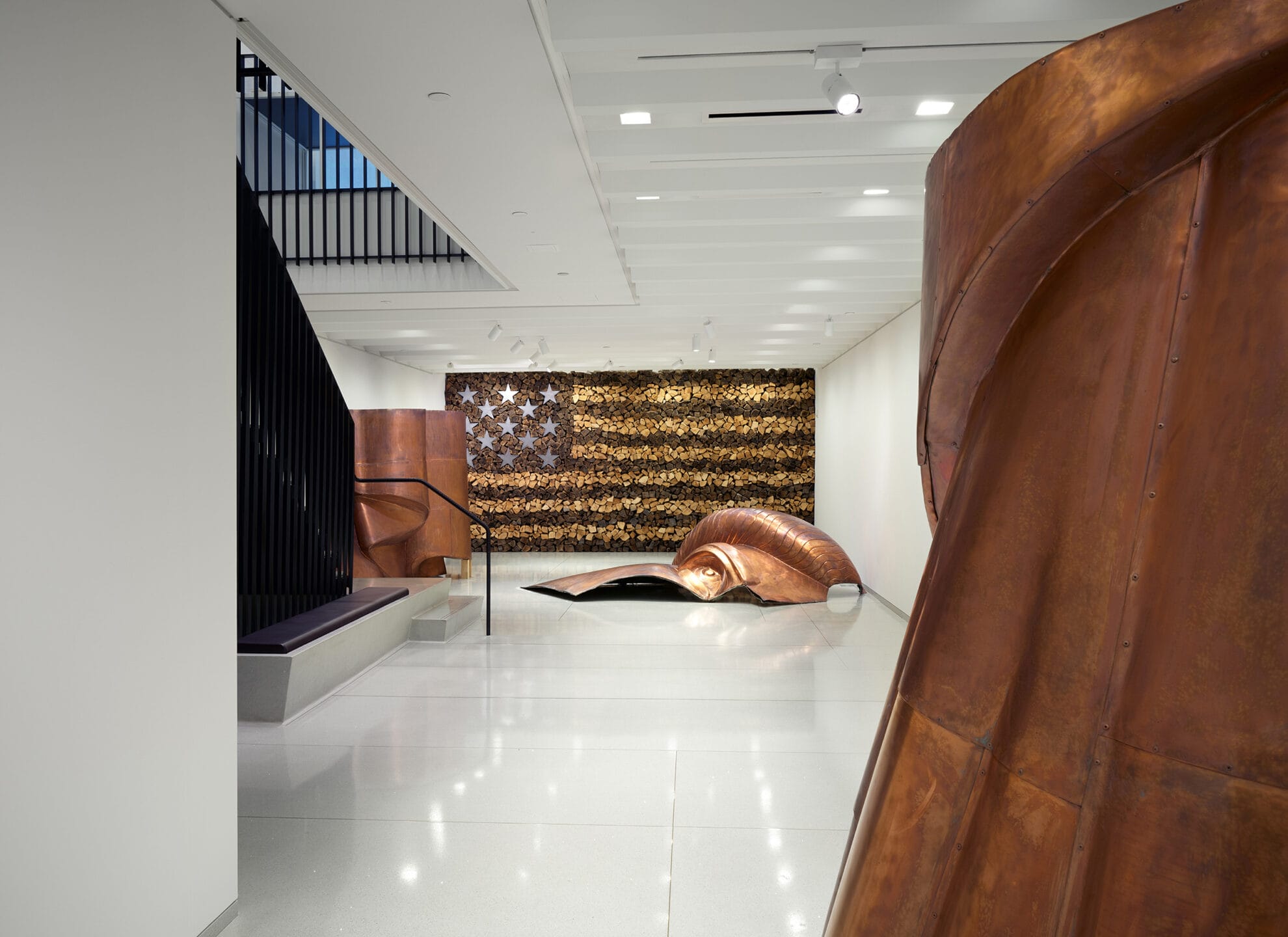 Modern art gallery featuring large copper sculptures and a wood log American flag wall installation.