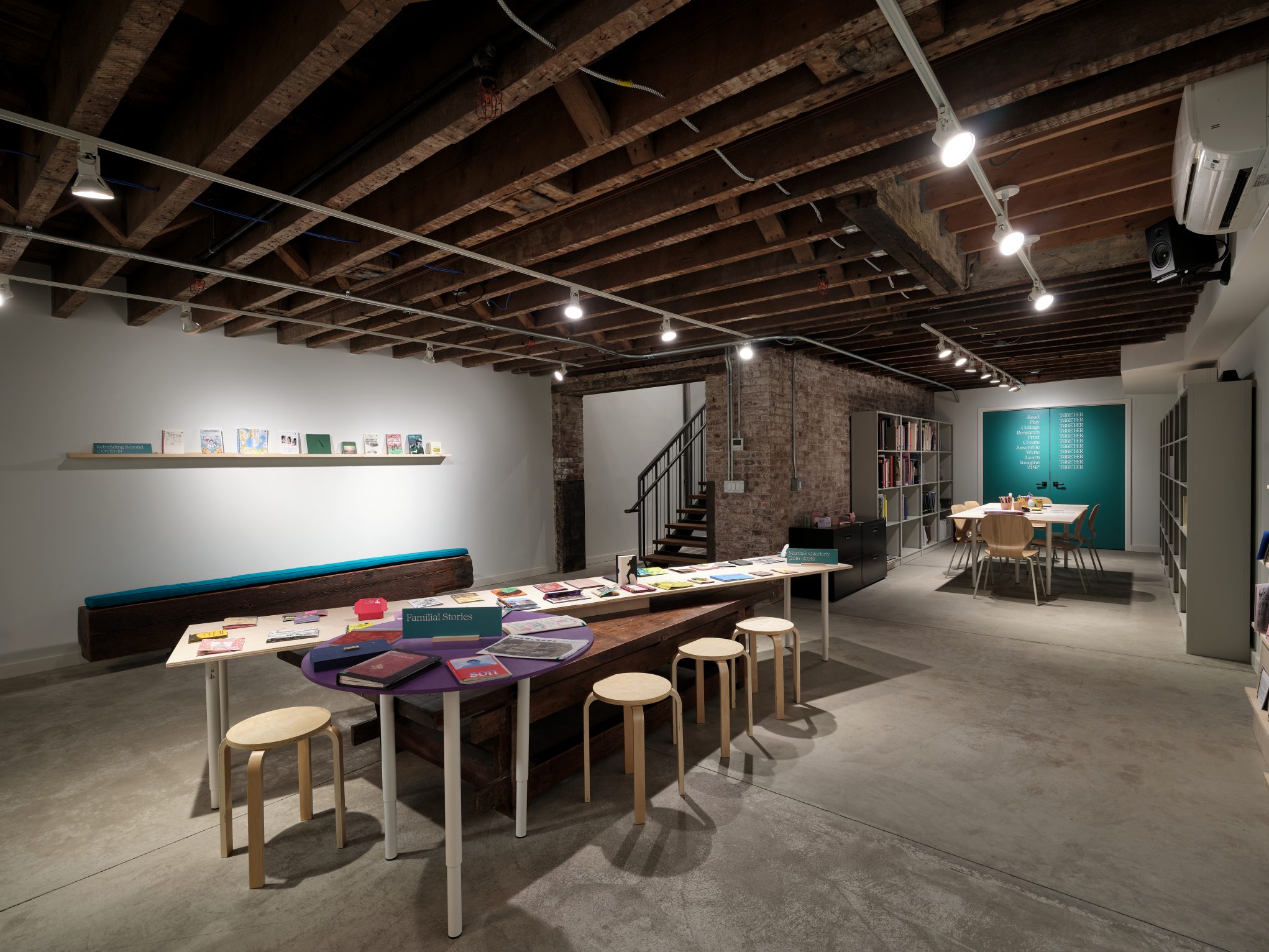 Industrial loft reading room with exposed beams, long tables and stools, bookshelves and zine display in creative studio.