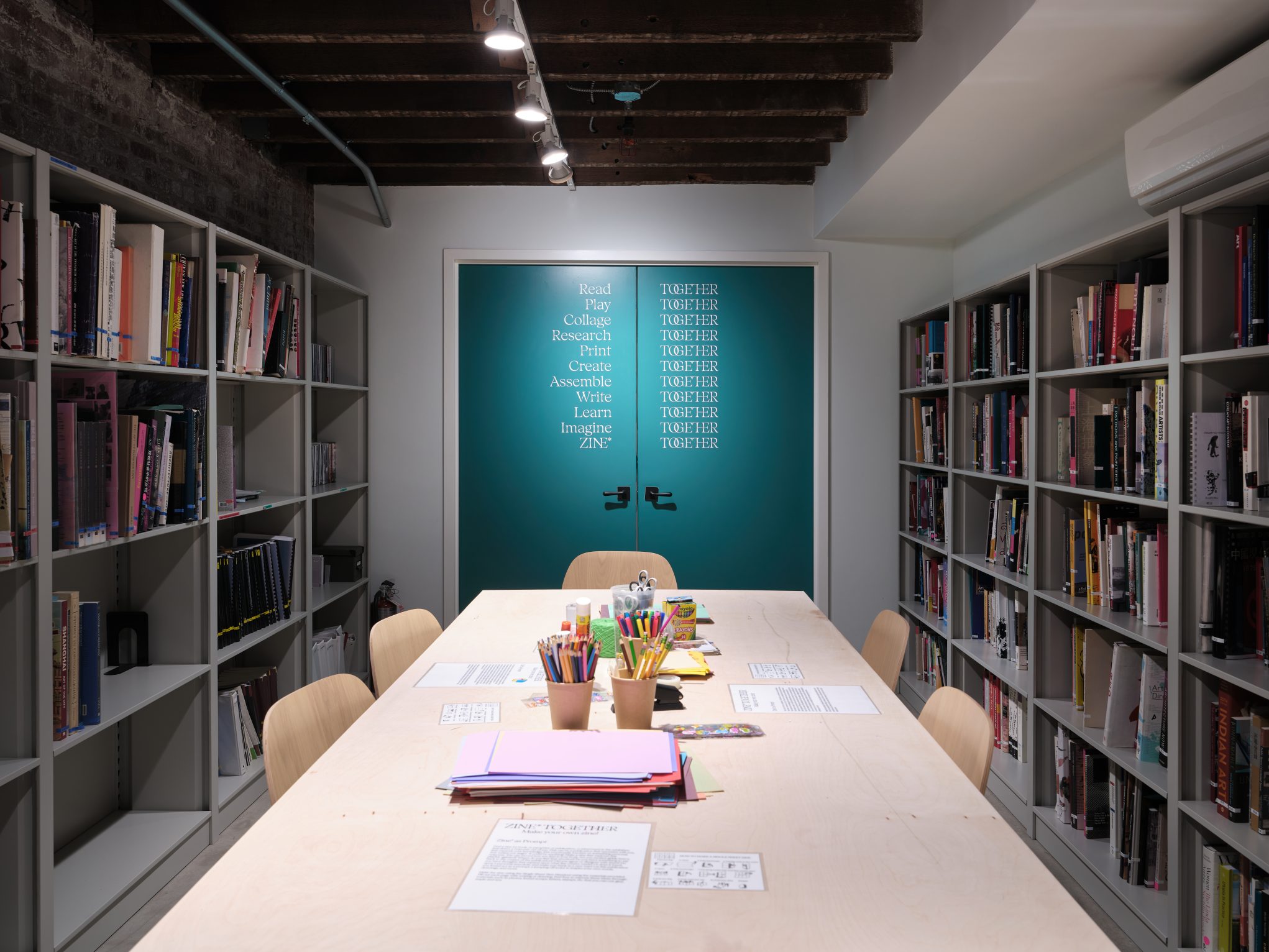 Library workshop room with long table, art supplies, and bookshelves, set up for a creative zine-making session.