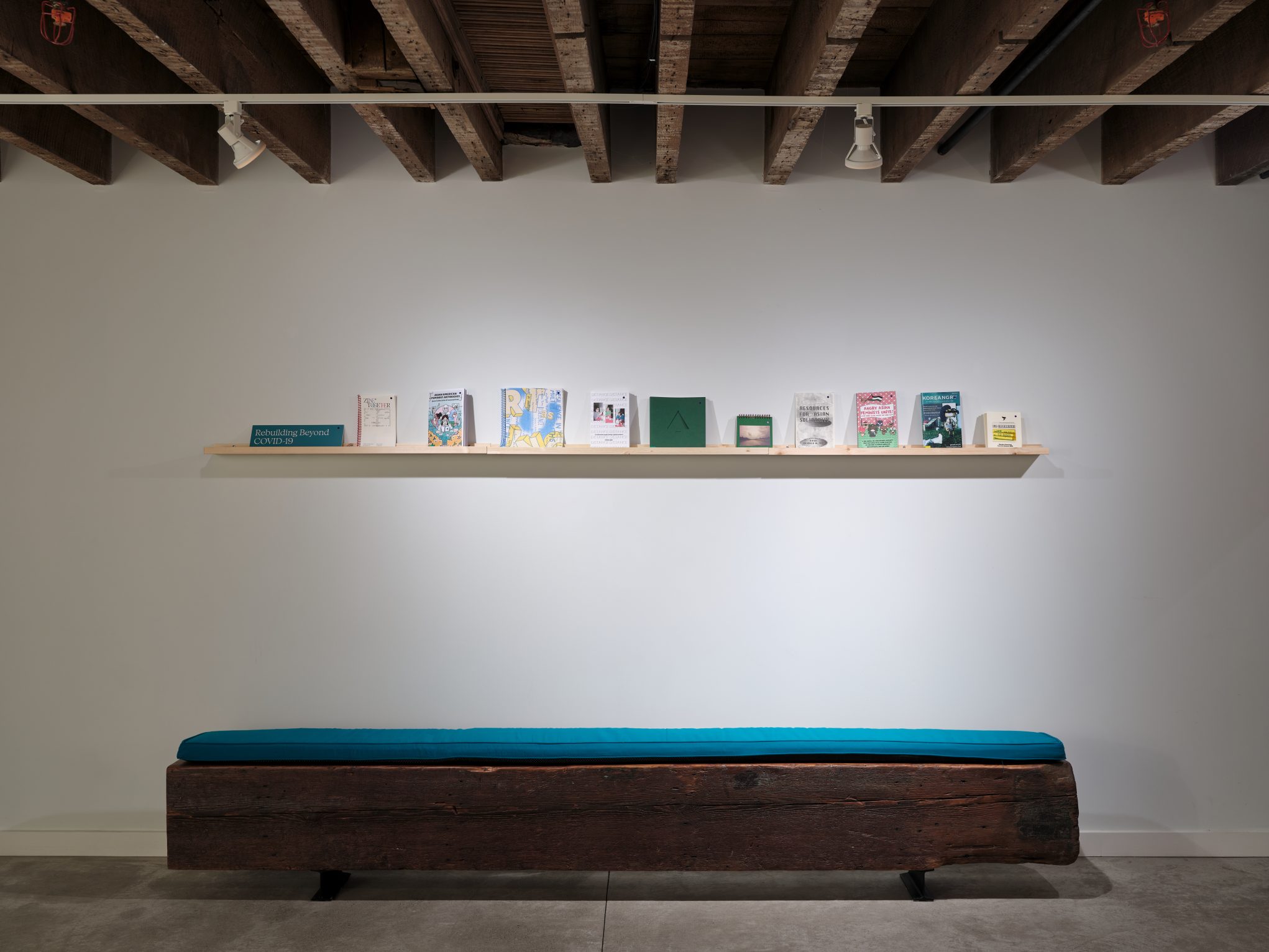 Minimalist gallery with rustic wood bench and teal cushion, floating shelf displaying books and zines under exposed beams.