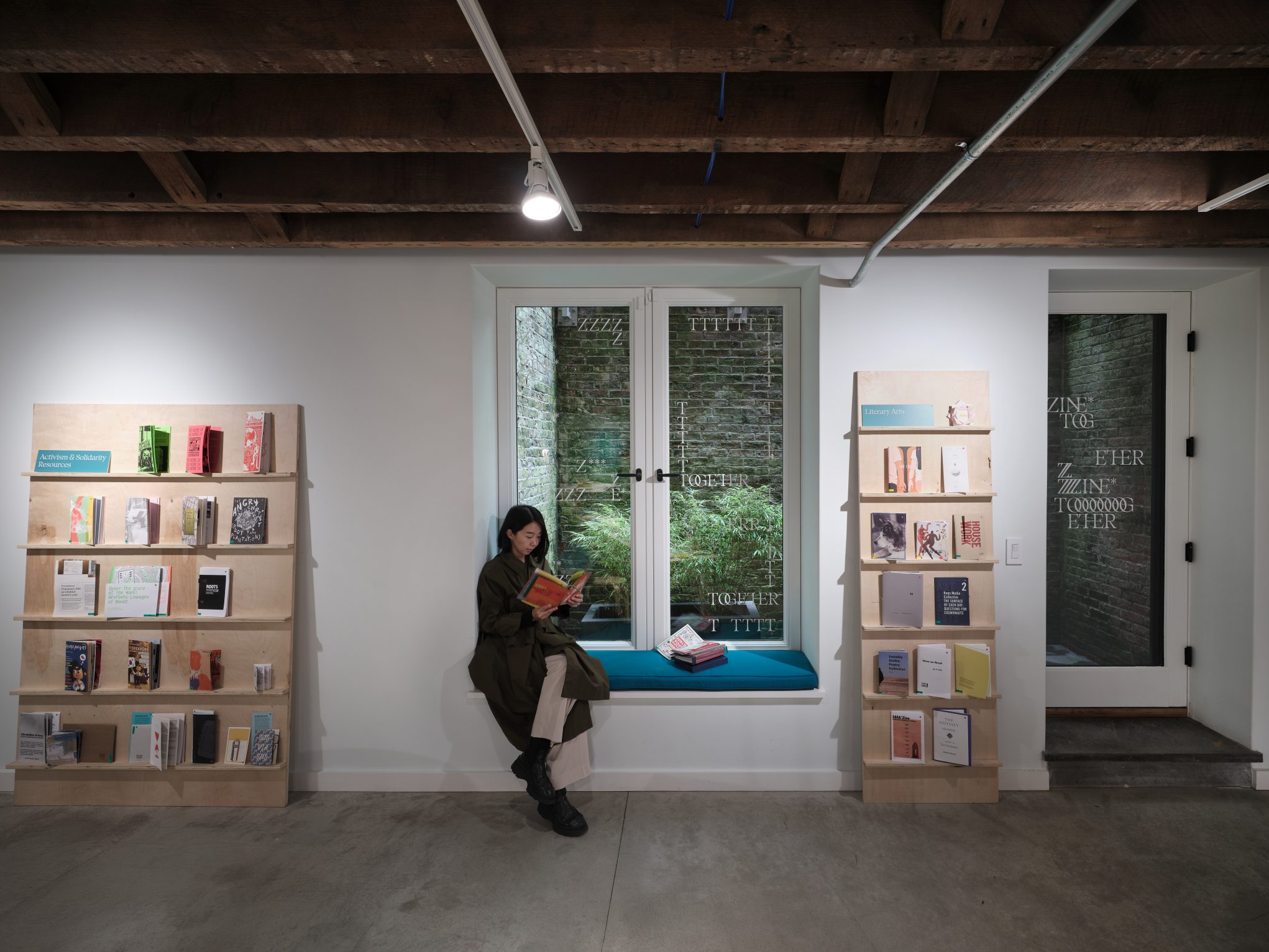 Modern bookstore with zine displays on wooden shelves; person reading at window seat, exposed beams, concrete floor.