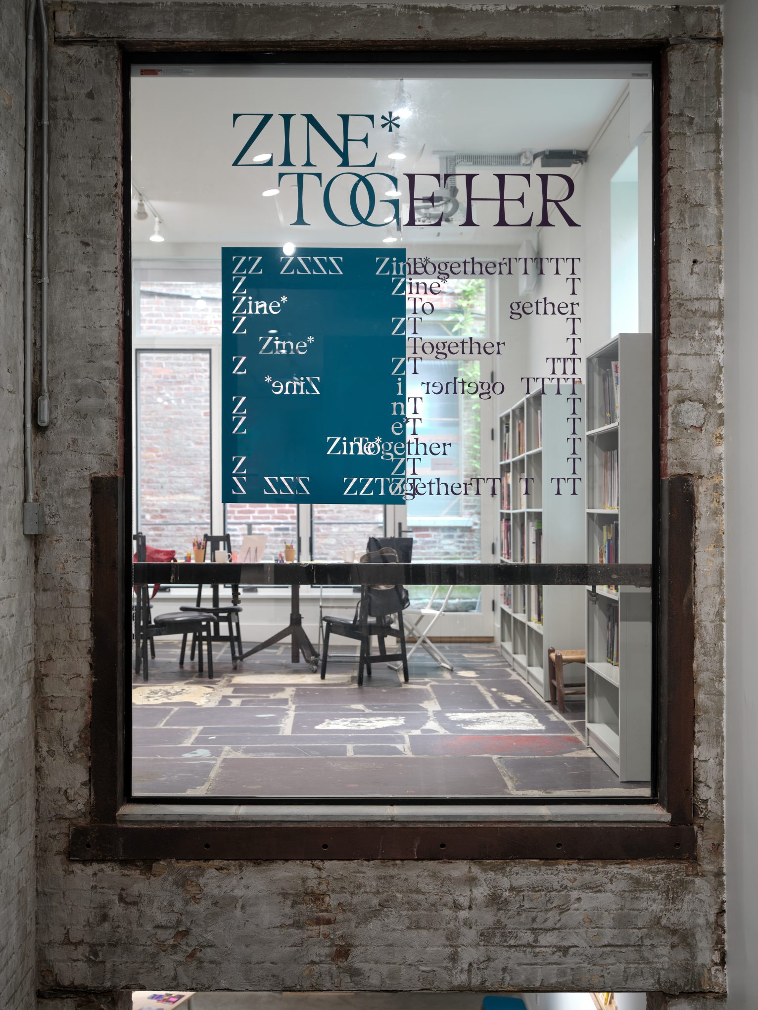 Zine Together studio and library seen through glass window with typographic decals, bookshelves, and chairs.