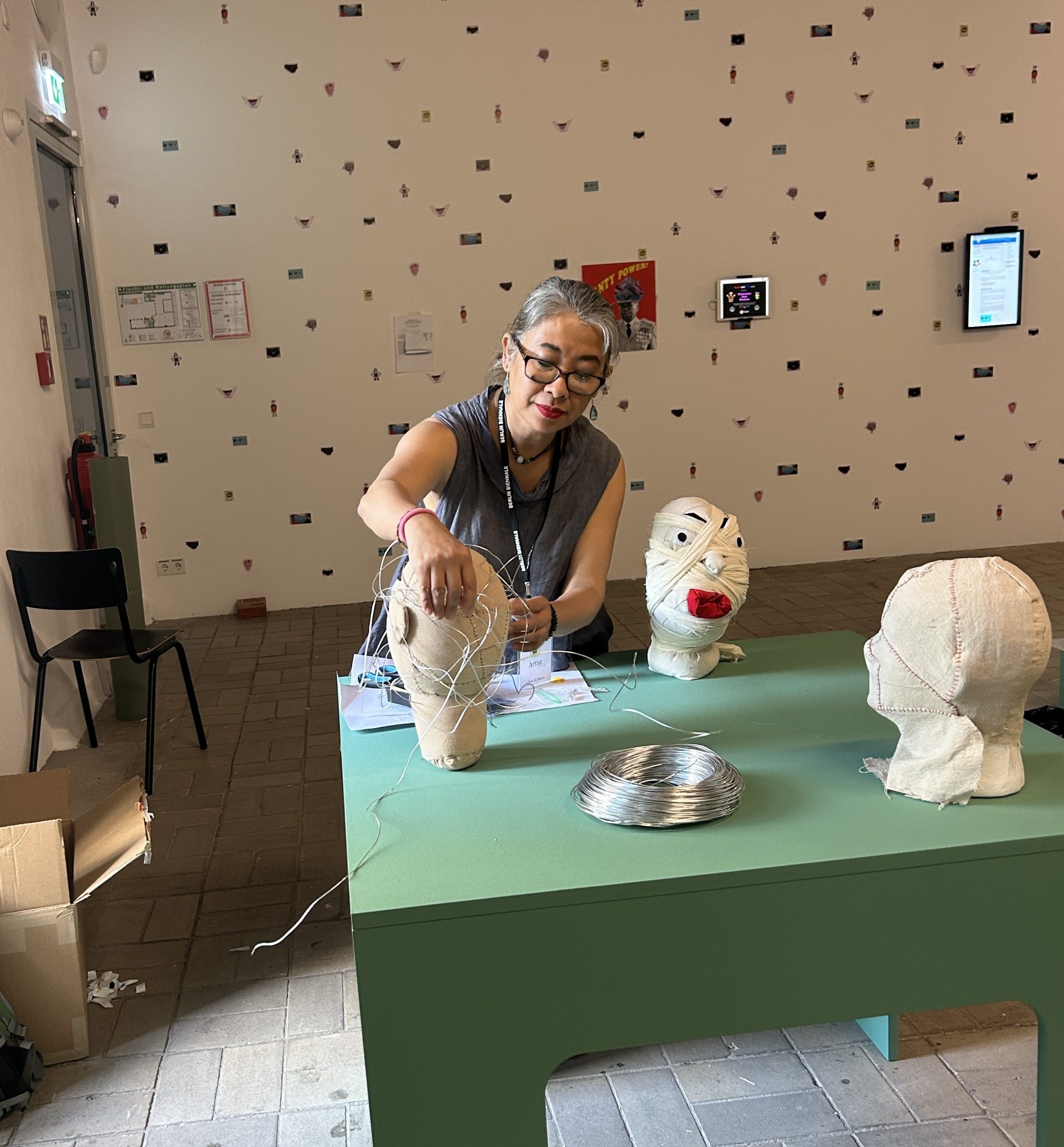 Artist crafting fabric-and-wire head sculptures at a gallery workshop table, modern art installation in background.