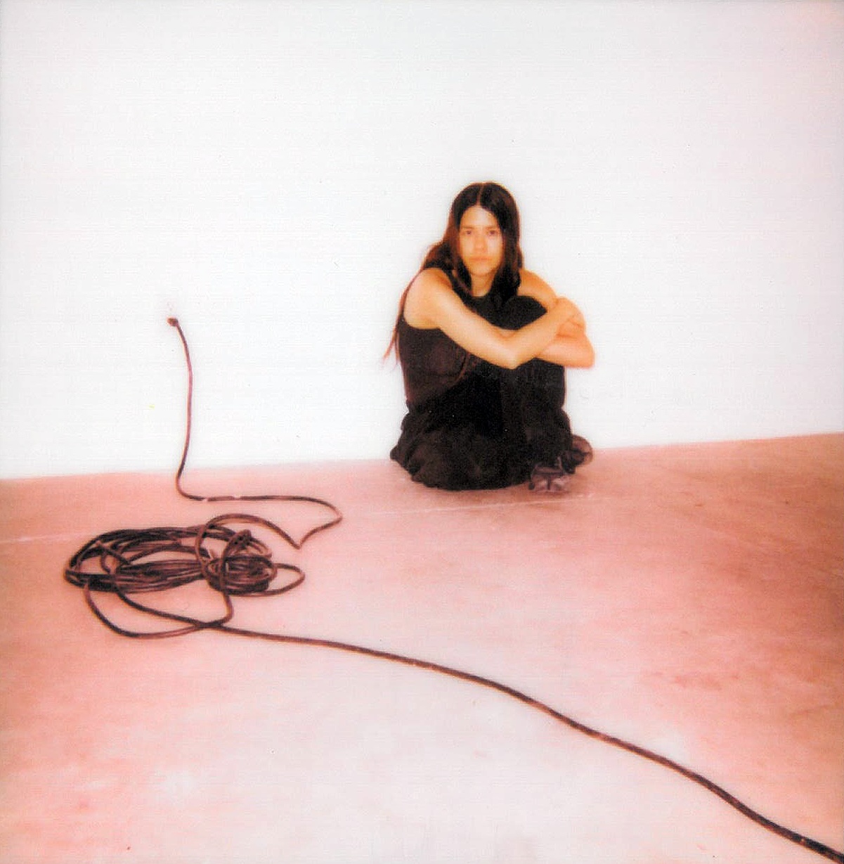 Minimalist studio portrait of person in black sitting on pink floor by white wall, coiled audio cable in foreground.