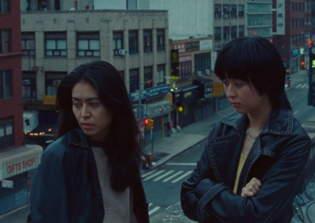 Two friends in leather jackets look pensive on a city rooftop overlooking Chinatown street at dusk.