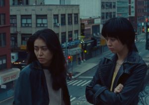 Two friends in leather jackets look pensive on a city rooftop overlooking Chinatown street at dusk.