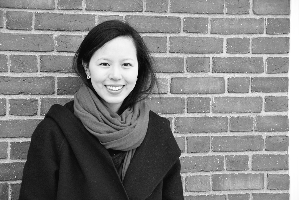 Black-and-white headshot of a smiling woman in coat and scarf against a brick wall background.