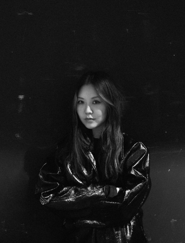 Black-and-white fashion portrait of a person in a shiny leather jacket, arms crossed, dark background, moody lighting.