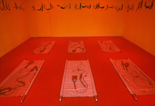 Contemporary art installation: six mesh cots on red floor, tangled hair and devices, orange gallery walls with calligraphy.