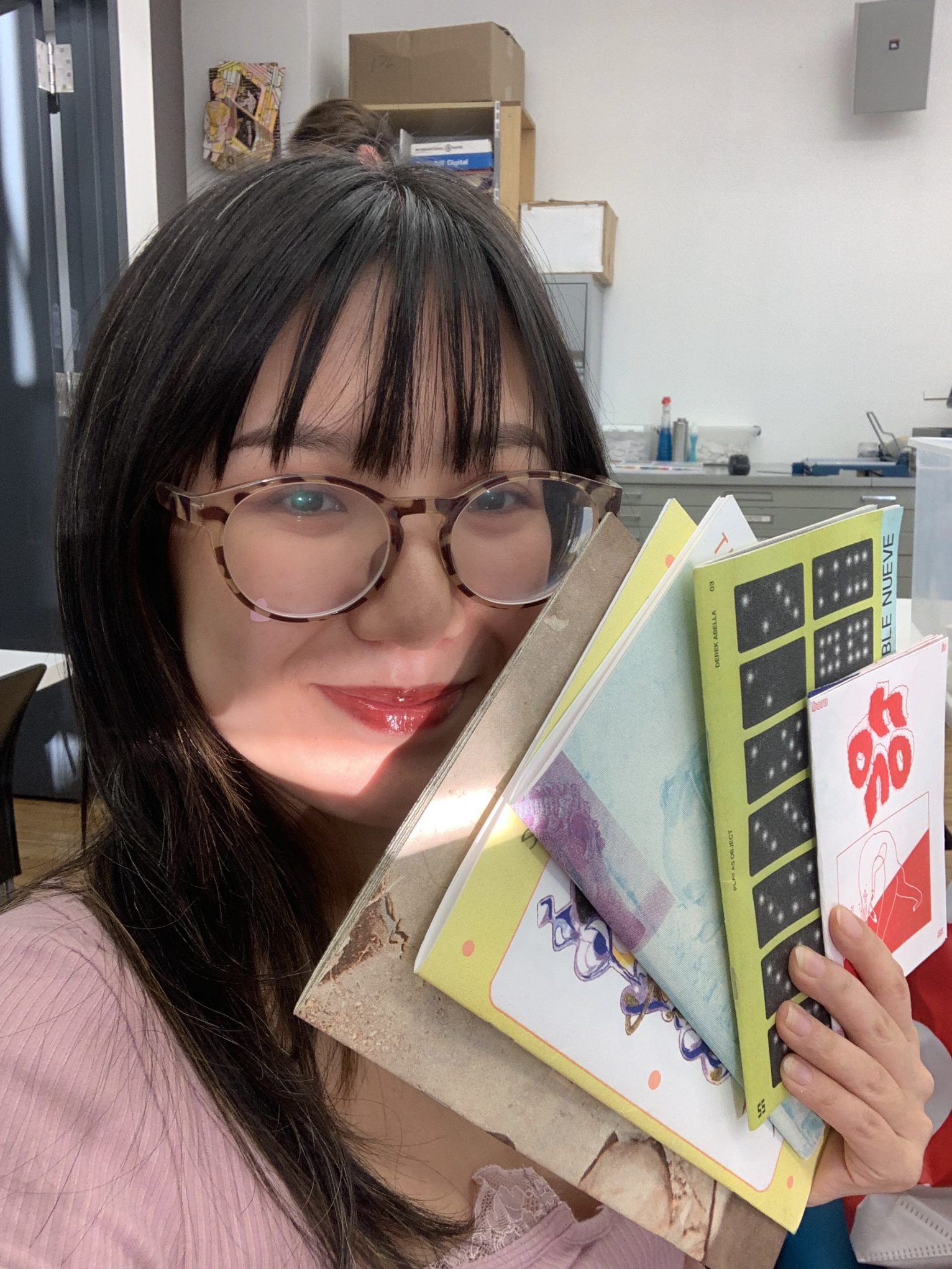 Selfie of smiling person with glasses holding colorful zines and art books in a bright creative studio.