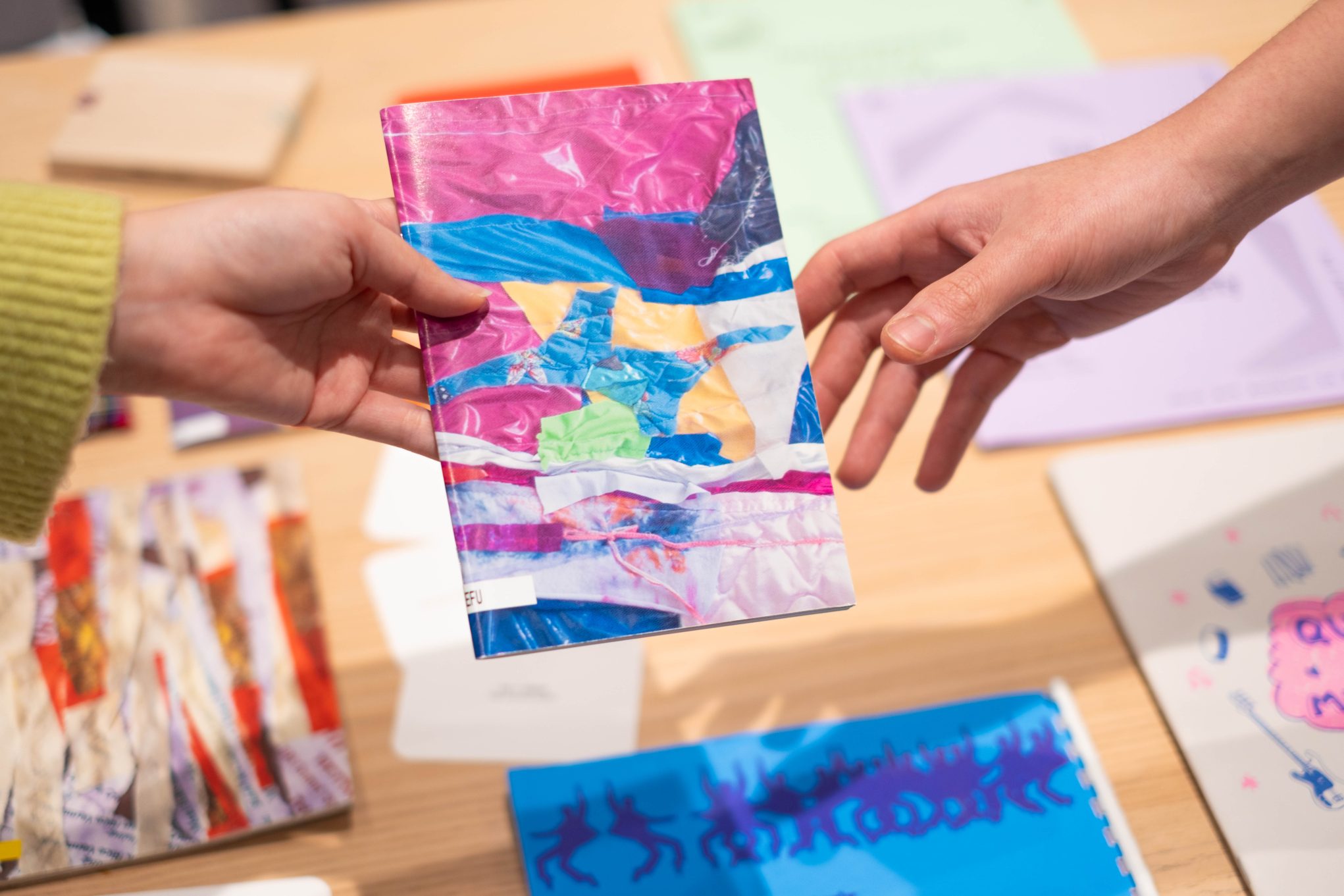 Hands exchanging a colorful handmade zine with abstract collage cover at an art fair table