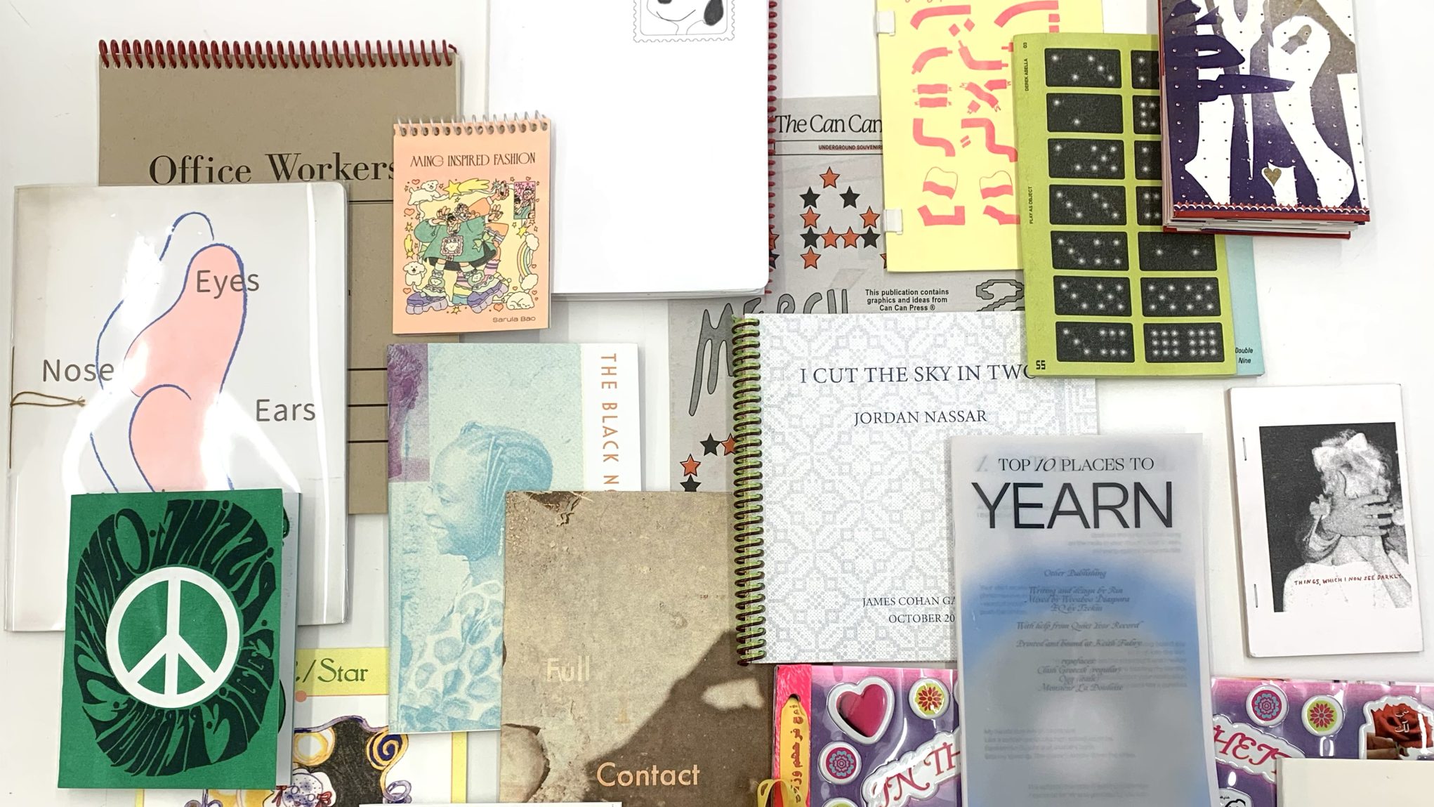 Flat lay of colorful indie zines, spiral notebooks, and art books arranged on a white background.