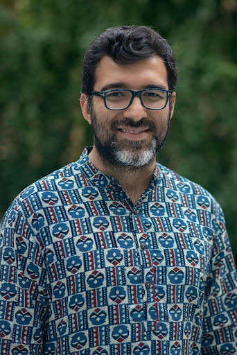Smiling bearded man with glasses in a blue patterned kurta-style shirt, outdoor portrait with green background.