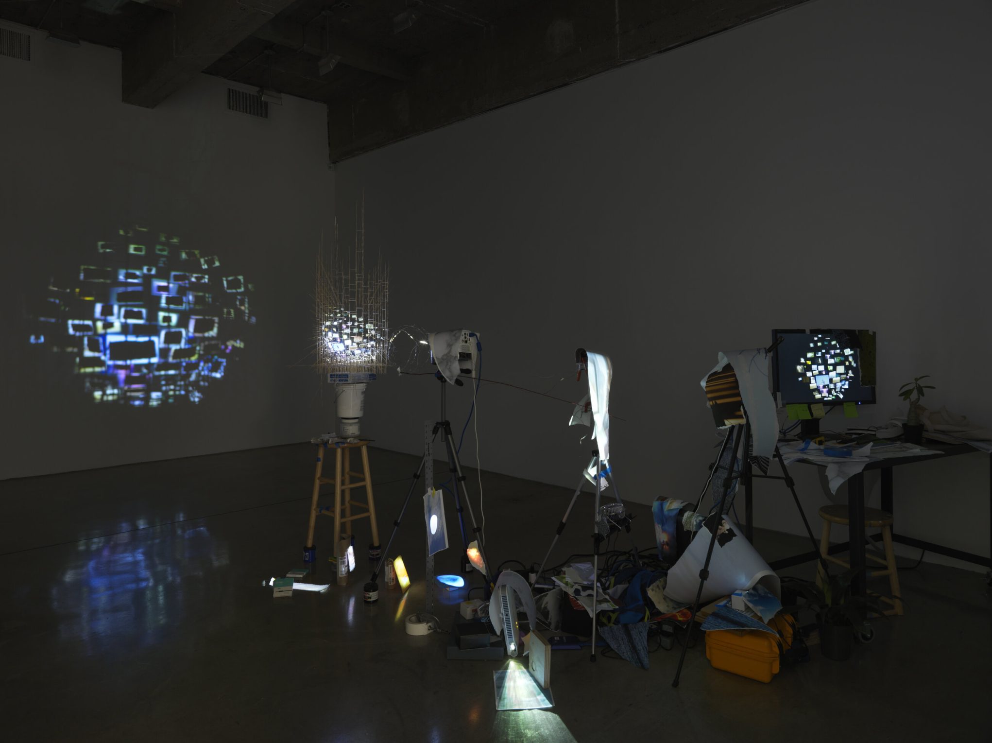 Mixed-media light projection art installation in dark gallery, tripods and monitors forming mosaic sphere visuals on wall.