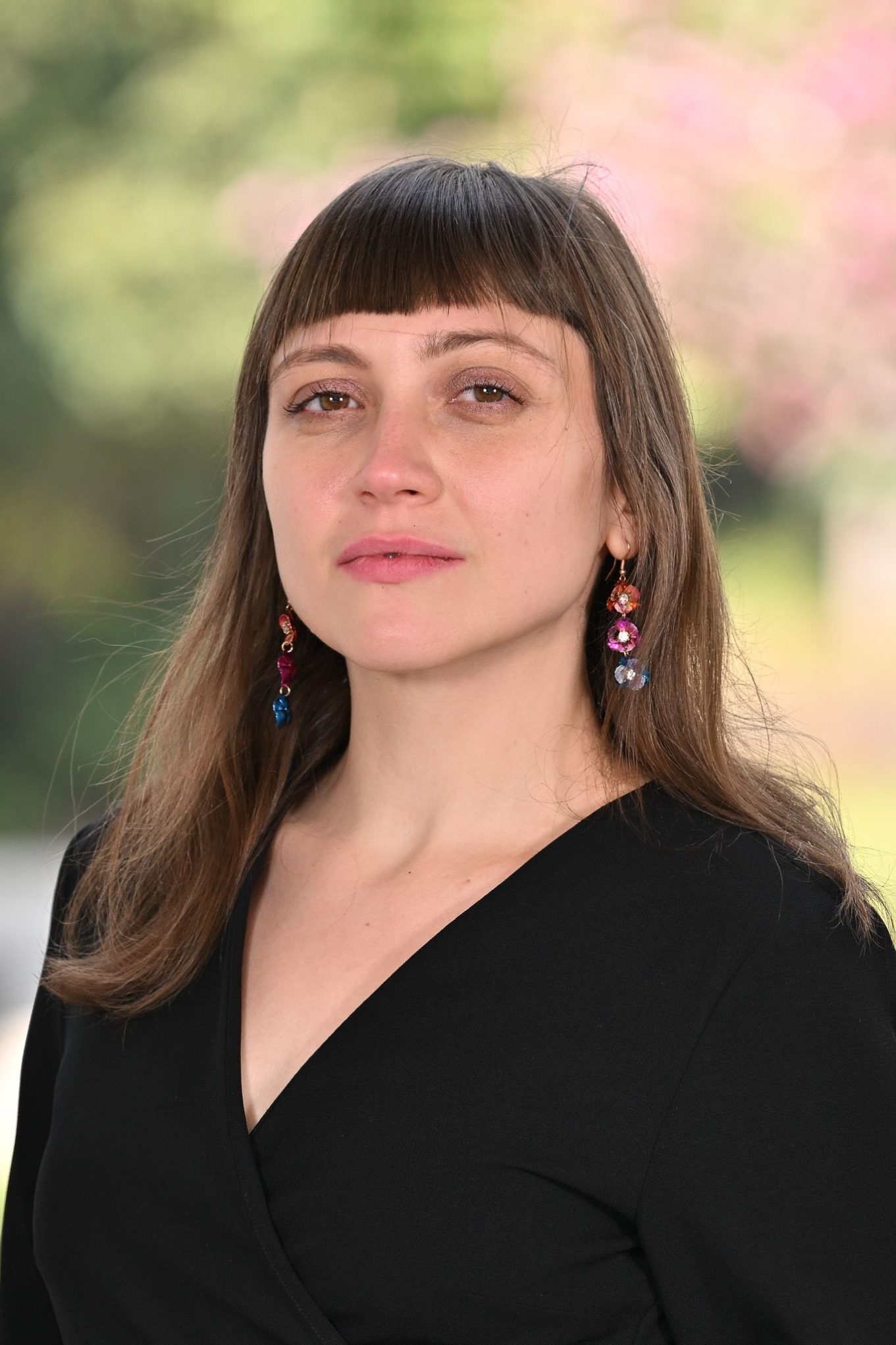 Professional headshot of woman with brown hair and bangs in black top and colorful earrings, outdoors blurred background