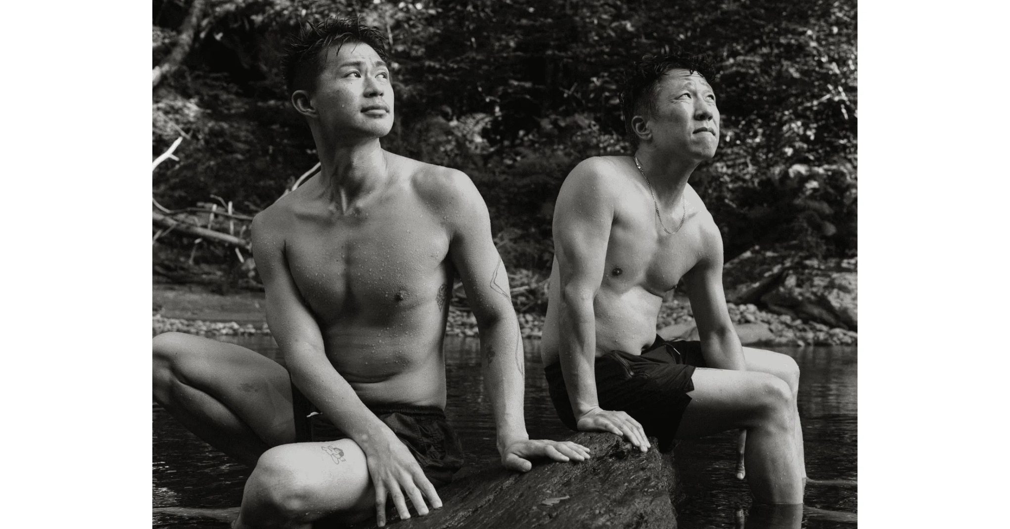 Black and white photo of two shirtless men sitting on a river rock in a forest, cooling off after a swim outdoors.