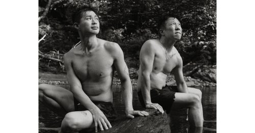 Black and white photo of two shirtless men sitting on a river rock in a forest, cooling off after a swim outdoors.