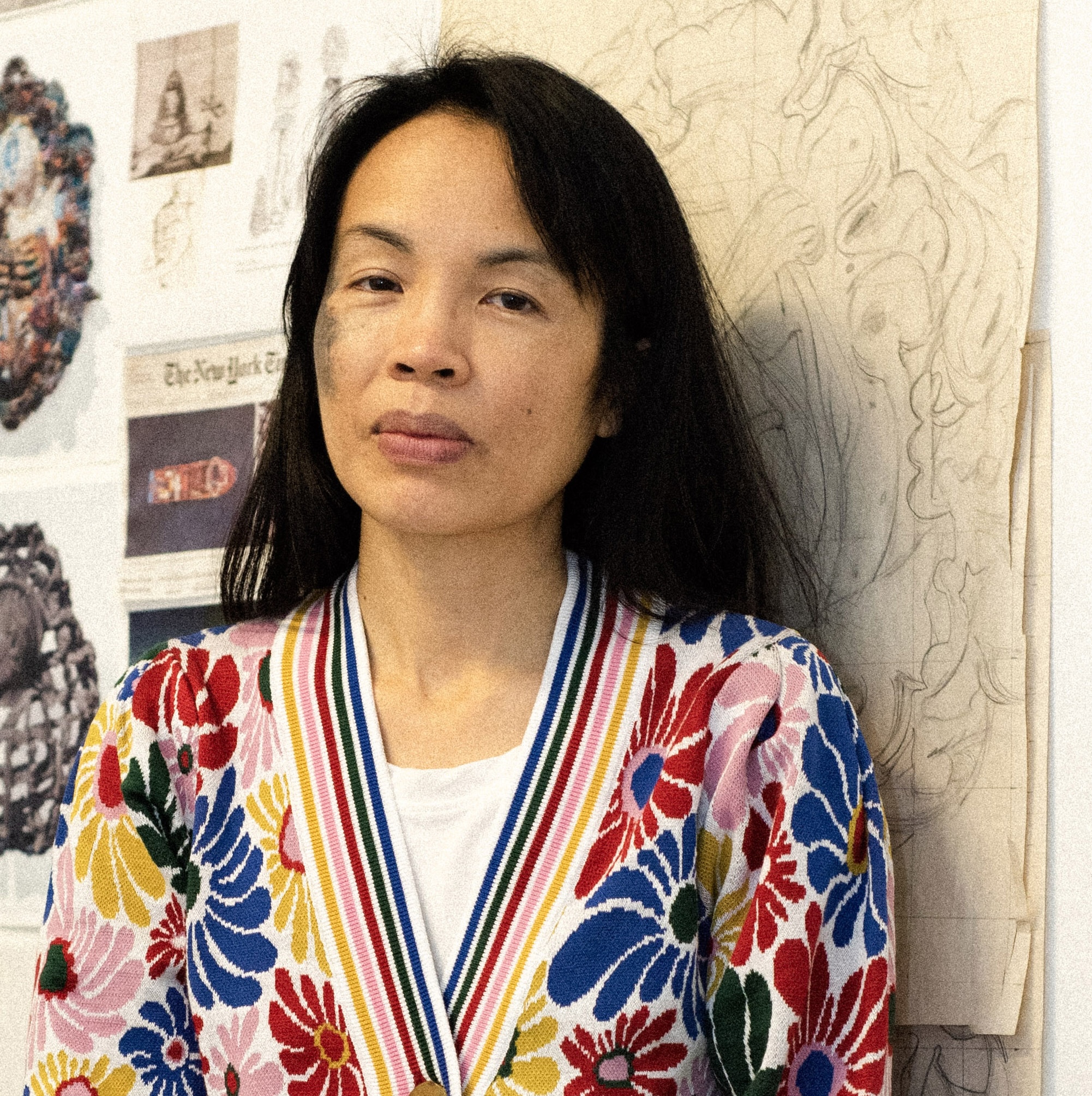 Portrait of an artist in a studio, wearing a colorful floral cardigan, standing before sketches and collage-covered wall.