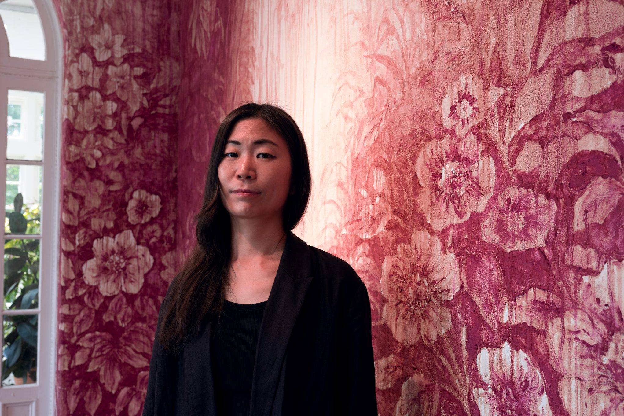 Woman in black blazer standing by pink floral mural in art gallery, soft window light