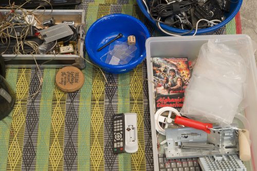 Yard sale assortment of electronics, cables, remotes, tools, and bins on a colorful woven mat