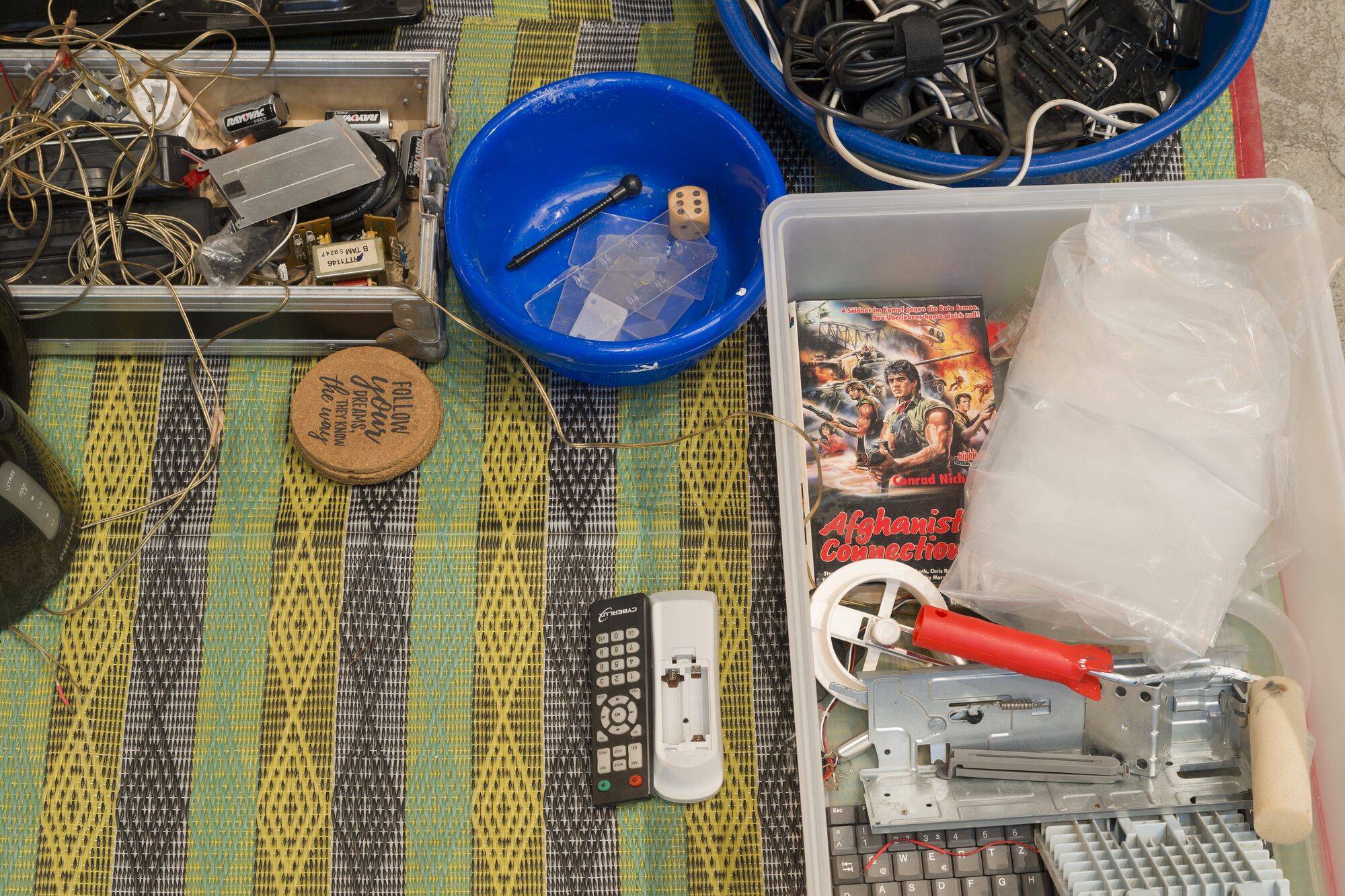 Yard sale assortment of electronics, cables, remotes, tools, and bins on a colorful woven mat
