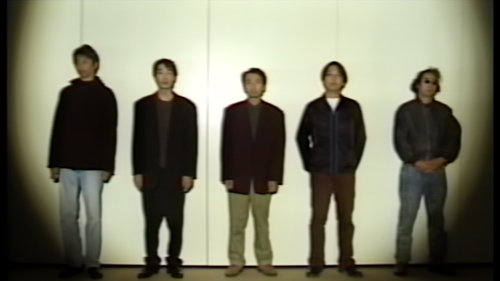 Five men stand in a lineup against a plain wall under a vignetted spotlight, wearing casual jackets and jeans, retro vibe.