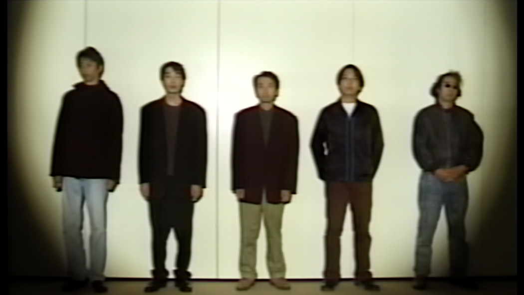 Five men stand in a lineup against a plain wall under a vignetted spotlight, wearing casual jackets and jeans, retro vibe.