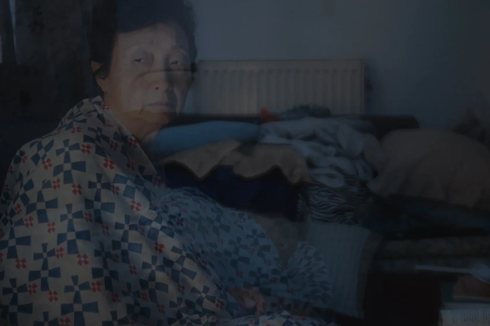 Elderly person in low light, wrapped in patterned blanket on messy bed, somber mood, night insomnia and loneliness.