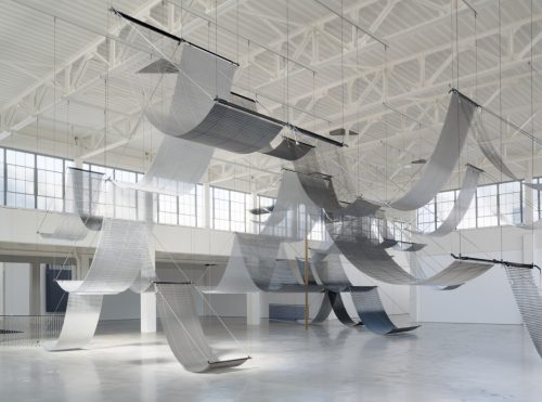Large-scale suspended metal mesh installation with draped waves in a contemporary art gallery space.