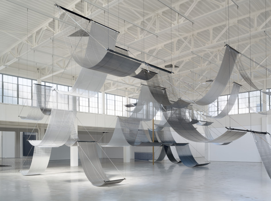 Large-scale suspended metal mesh installation with draped waves in a contemporary art gallery space.