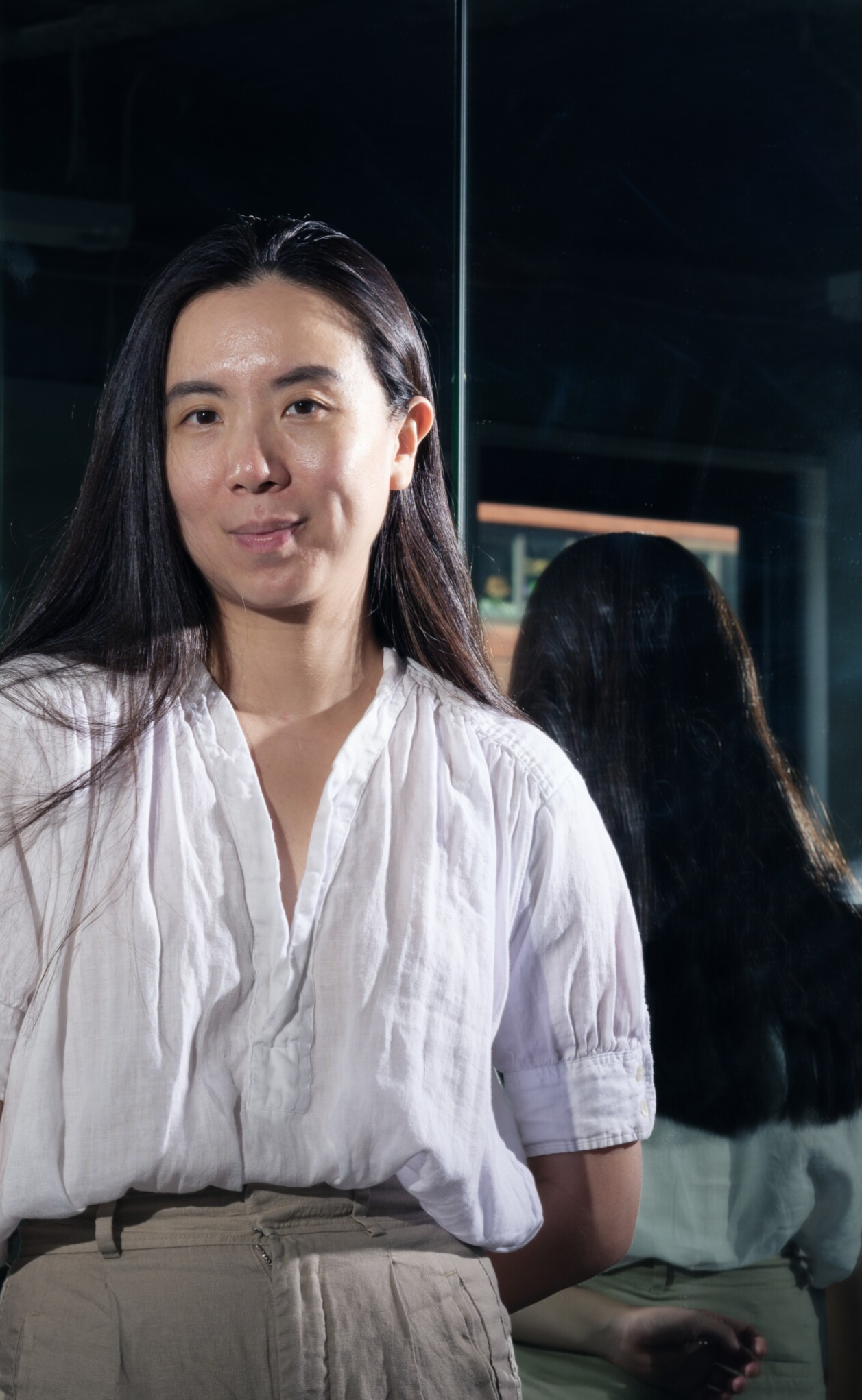 Portrait of a woman in a white blouse with long hair, standing by a mirror with reflection, indoor studio lighting.